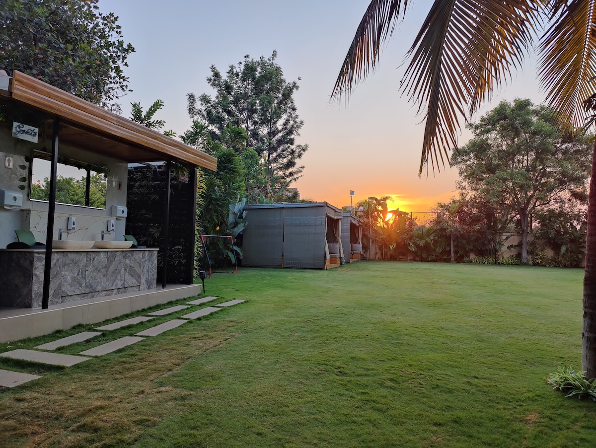 A well-maintained garden area is featured with lush green grass leading to a serene sunset. A stone pathway is visible alongside a stylish outdoor space with wash basins, framed by tropical trees and cabinets, providing a peaceful atmosphere.