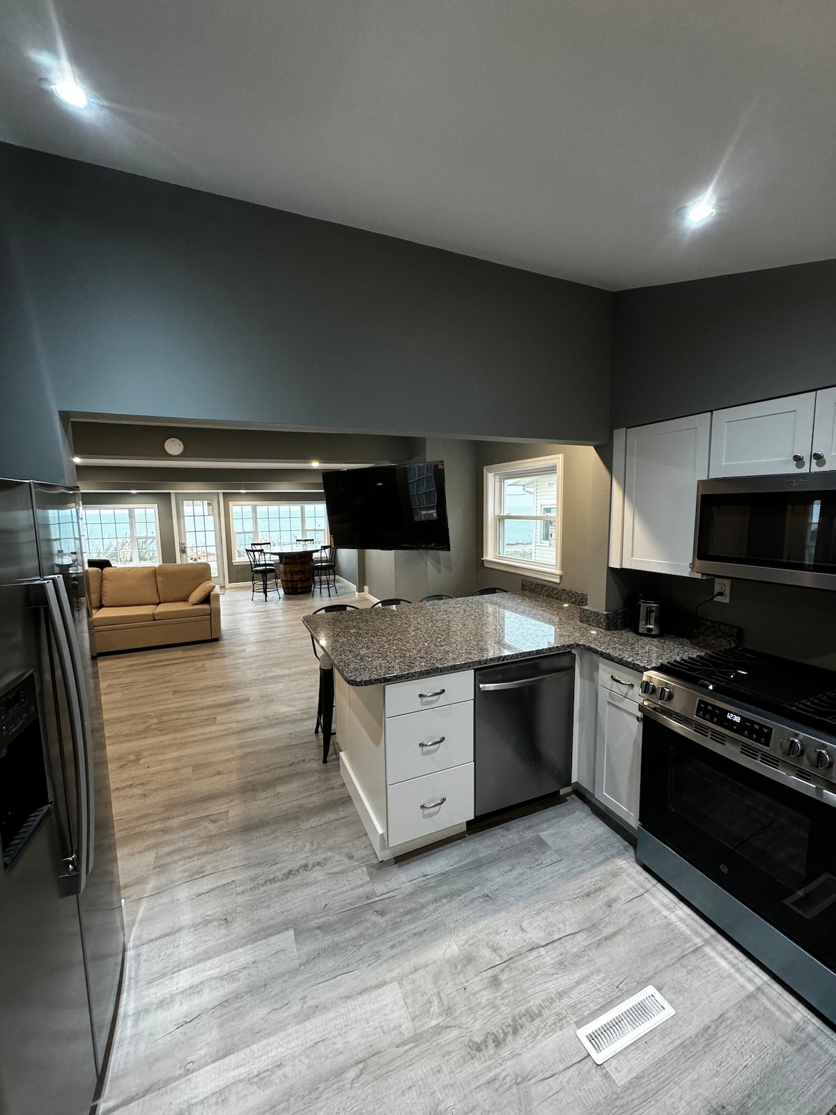 The kitchen is equipped with modern appliances and features a spacious granite island. Light grey cabinetry complements the sleek design. A view of an adjoining living area with natural light is visible through large windows, providing a welcoming connection to shared spaces.