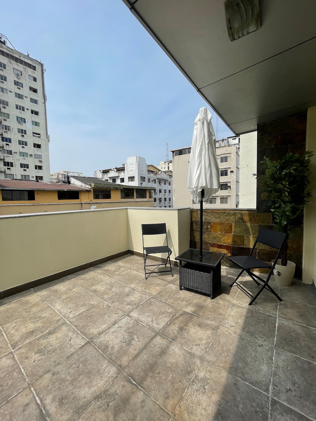 A private terrace features tiled flooring and is furnished with two black folding chairs and a small table. A white umbrella provides shade, and a potted plant adds a touch of greenery. Surrounding buildings create an urban backdrop.