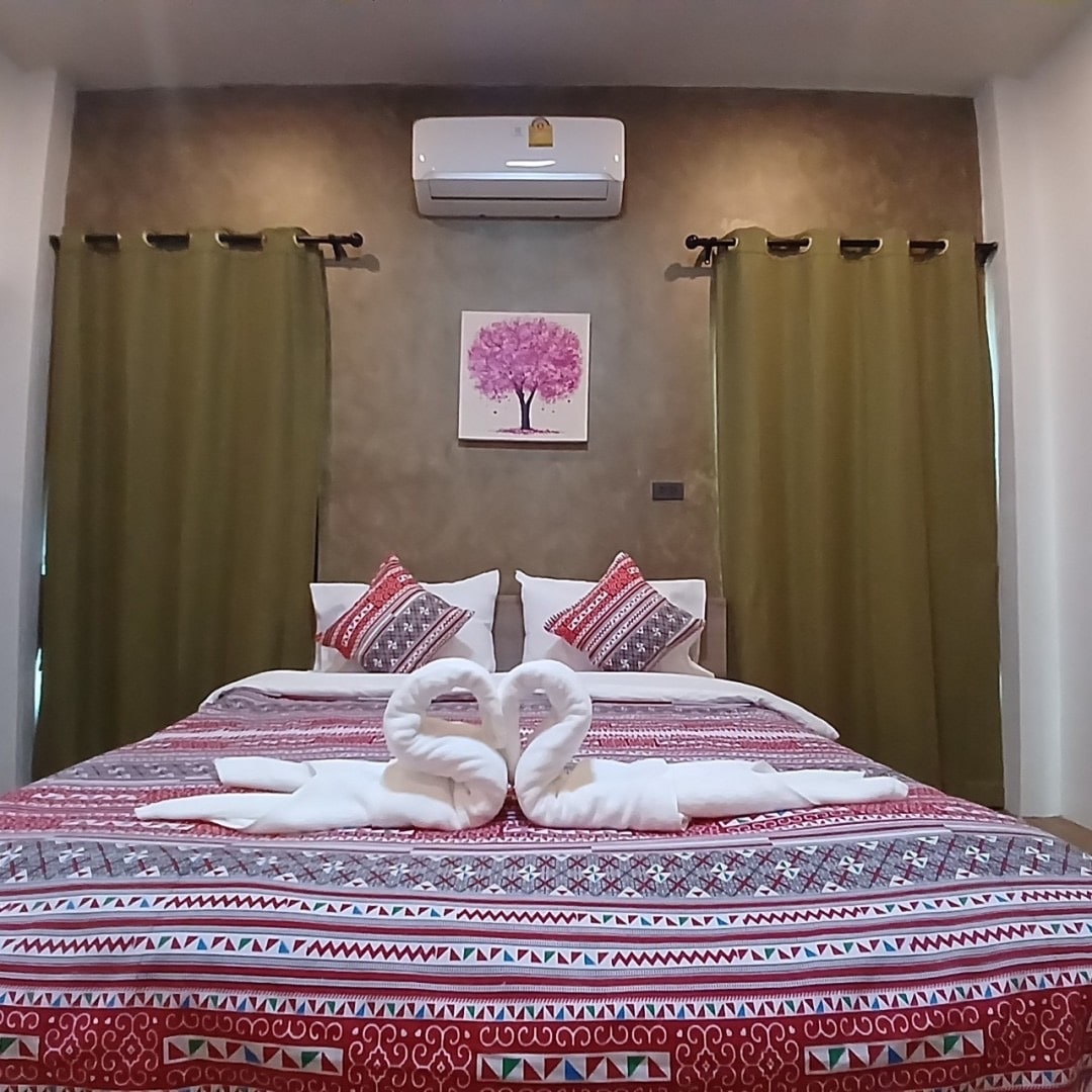 A comfortable sleeping area features a spacious bed adorned with decorative pillows. Soft curtains frame the windows, and a wall-mounted air conditioning unit is positioned above. A vibrant artwork depicting a tree adds a pop of color to the neutral wall backdrop.