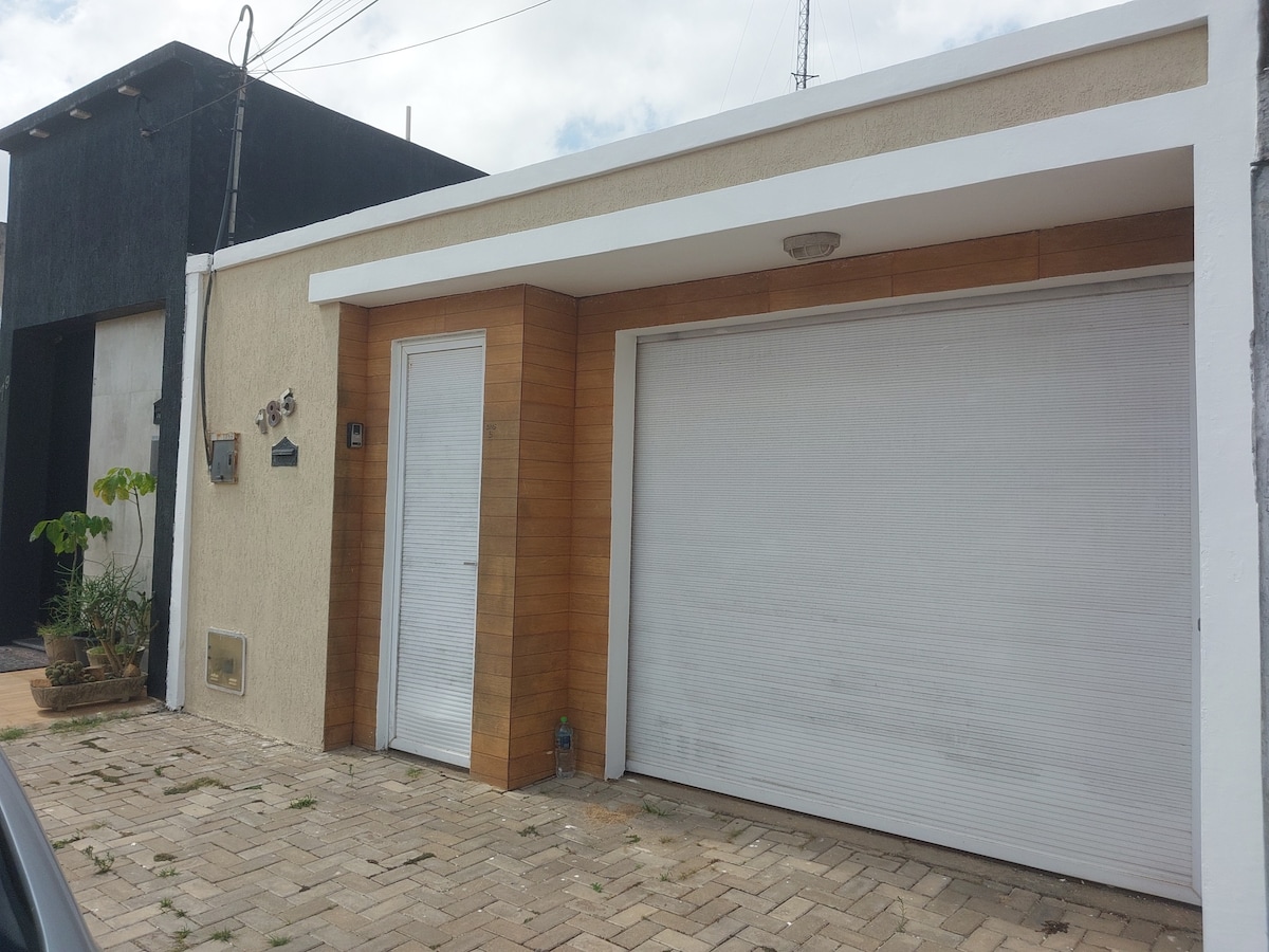 The exterior of a single-story building features a combination of light-colored stucco and wooden accents. A large closed garage door is present, along with a pedestrian entrance. Nearby, a small plant stands in a pot on the paved pathway.