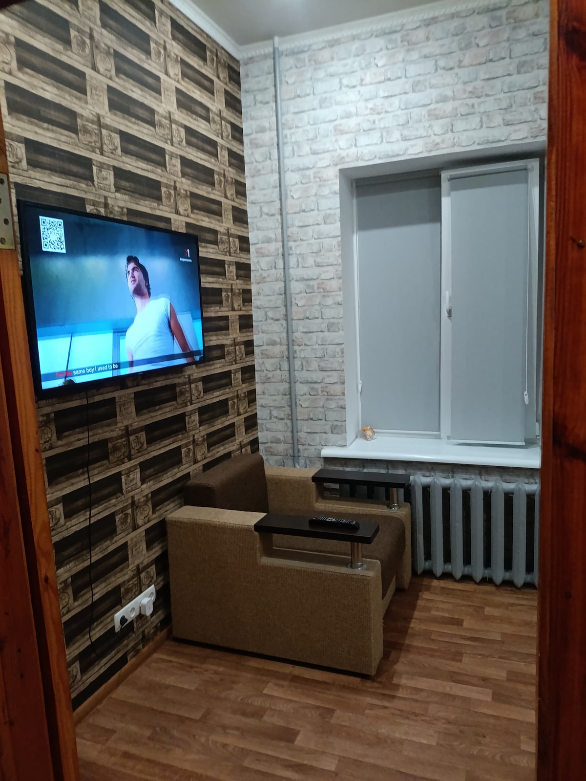 A small seating area is positioned next to a window, featuring a fabric armchair with a tiny side table. A wall-mounted television is visible, with a decorative wallpaper providing visual interest. Natural light streams in through the partially open blinds.