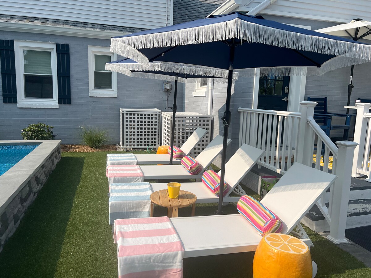 Three lounge chairs with colorful striped cushions are arranged by a heated pool. A small wooden table and a bright yellow accessory add a playful touch. Shade is provided by two blue and white umbrellas, while green grass surrounds the area for a refreshing outdoor space.
