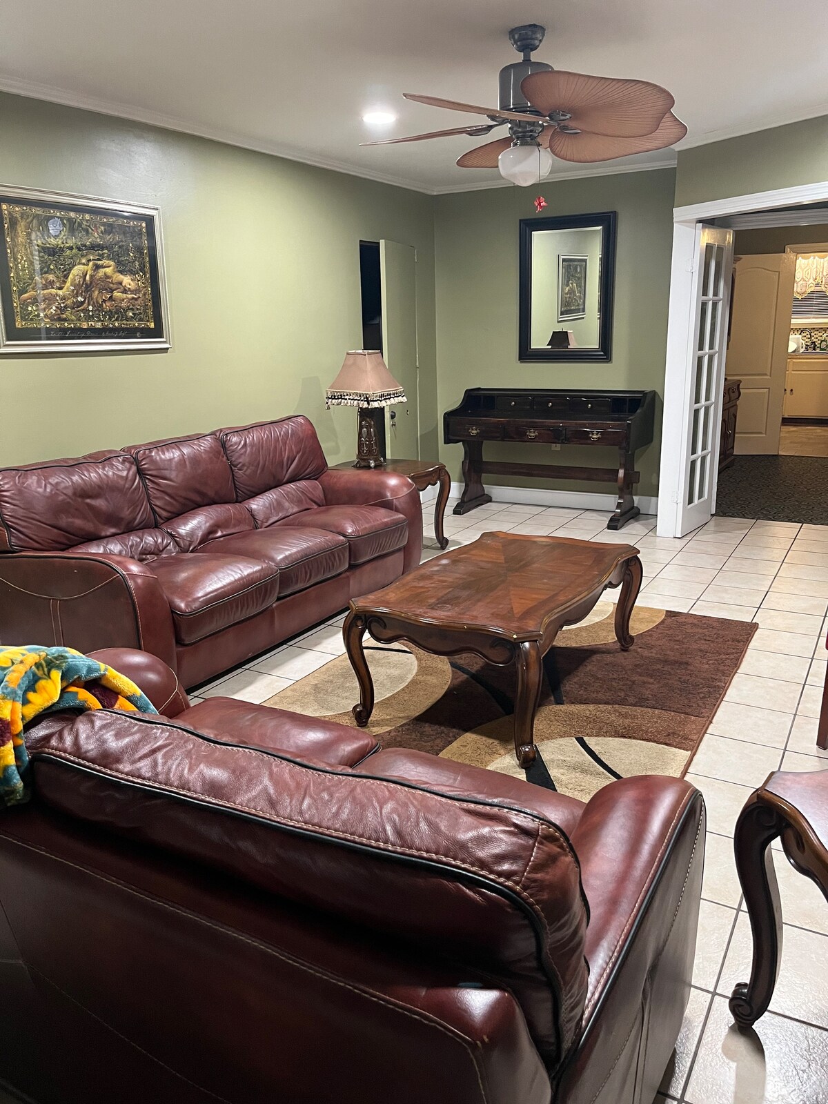A living area is presented with leather sofas and a coffee table atop a decorative rug. A lamp and framed artwork add to the decor, while natural light enters through open french doors leading to another room.