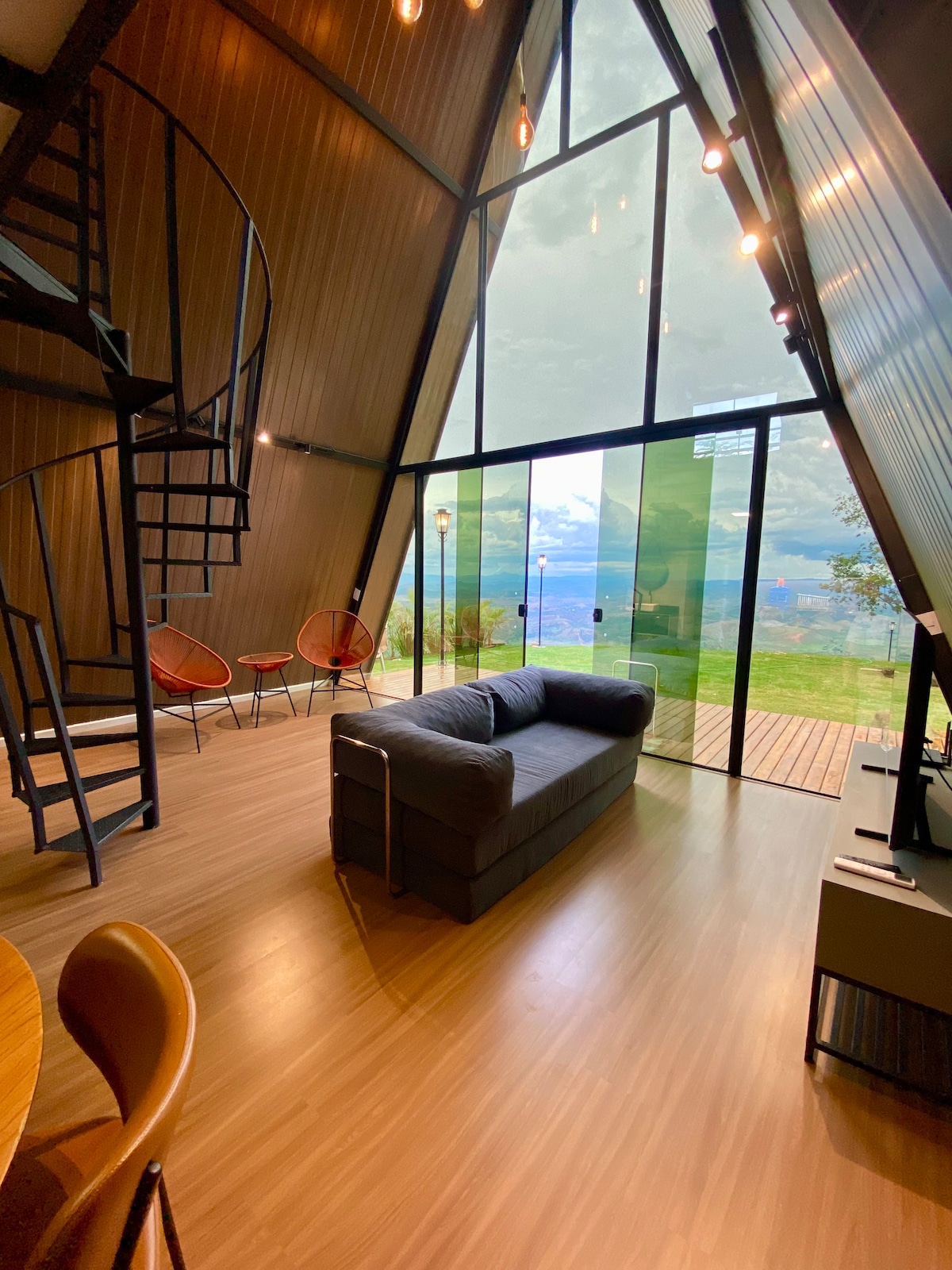 A modern interior presents a spiral staircase leading to an upper level. Large glass panels allow natural light to fill the space, offering a view of the lush surroundings. A sectional sofa is positioned for relaxation, while two orange chairs are set beside a round table.