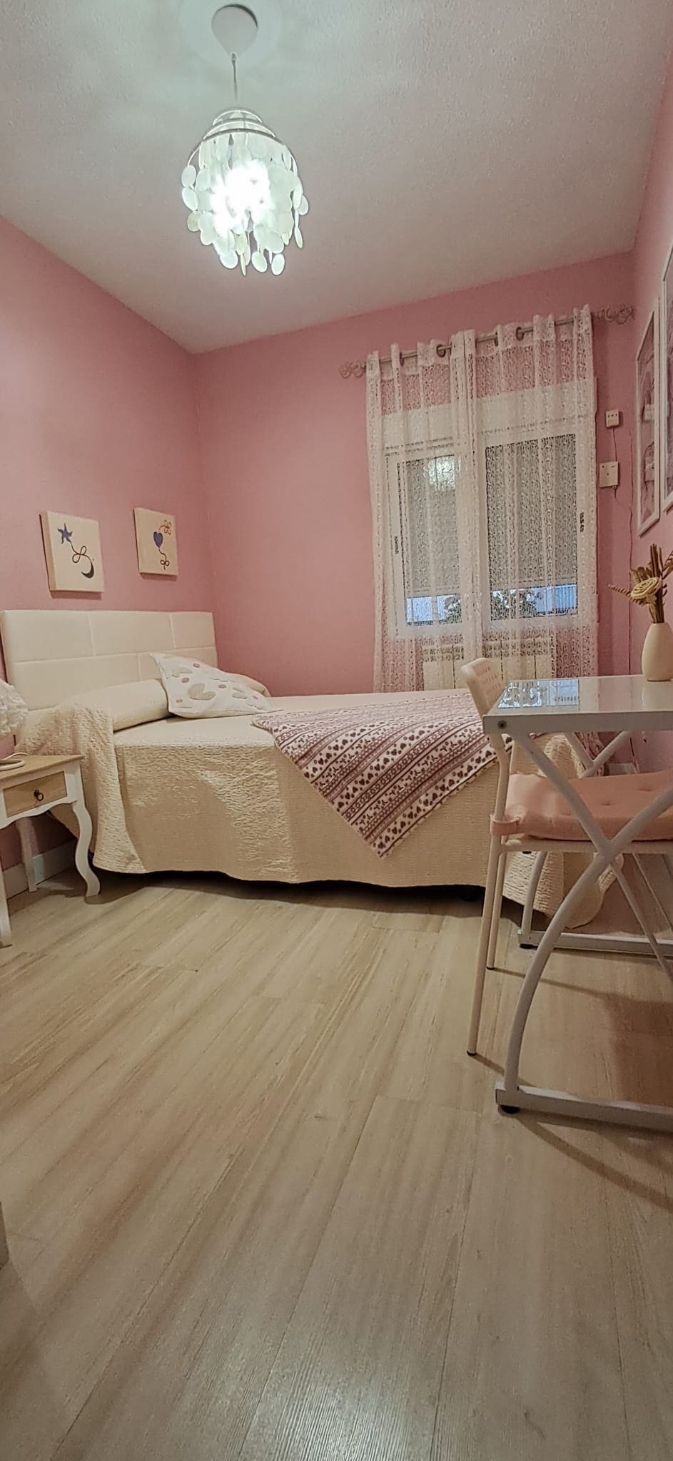 A cozy bedroom features soft pink walls and light wood flooring. A spacious bed is dressed in neutral tones, while two framed artworks adorn the wall above. A small desk is positioned near a window, allowing for natural light to brighten the space.