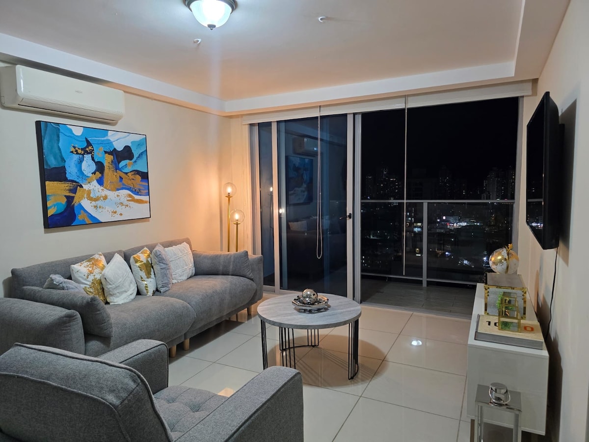 A living room features a cozy sofa and chair, positioned around a circular table. A painting with vibrant colors adorns the wall, while large glass windows offer a view of the city at night. Soft lighting provides a warm ambiance.