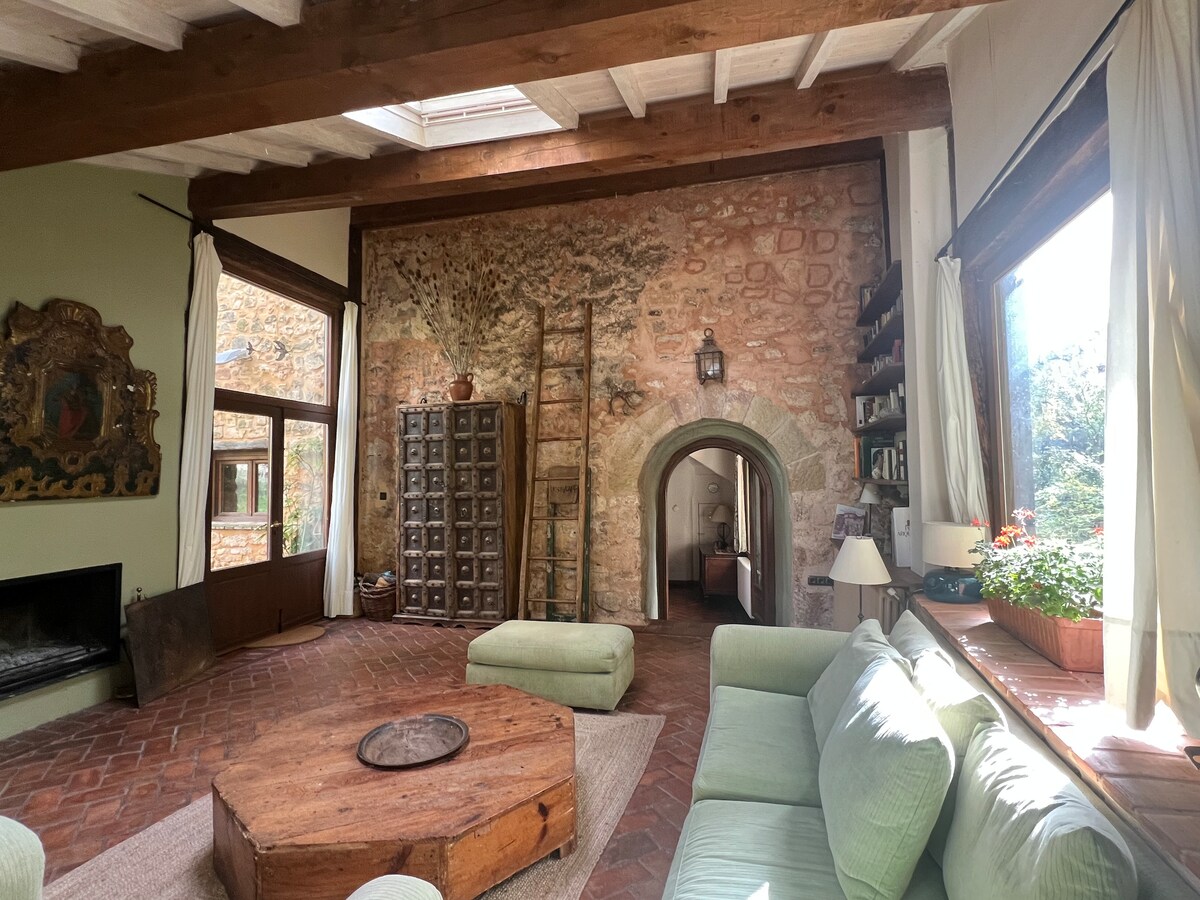 A spacious living area features rustic stone walls and large windows allowing natural light to illuminate the room. A round wooden coffee table is centered on the terracotta tiled floor, surrounded by soft seating in light colors. Decor incorporates natural elements, enhancing the serene ambiance.