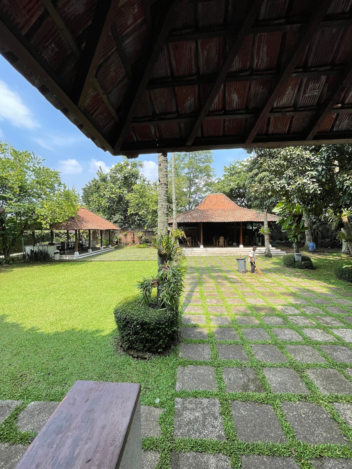 An expansive garden area showcases well-maintained grassy lawns bordered by neatly arranged stone paths. Traditional-style buildings with sloped roofs are visible in the background, surrounded by various trees and plants, creating a serene outdoor environment illuminated by natural light.