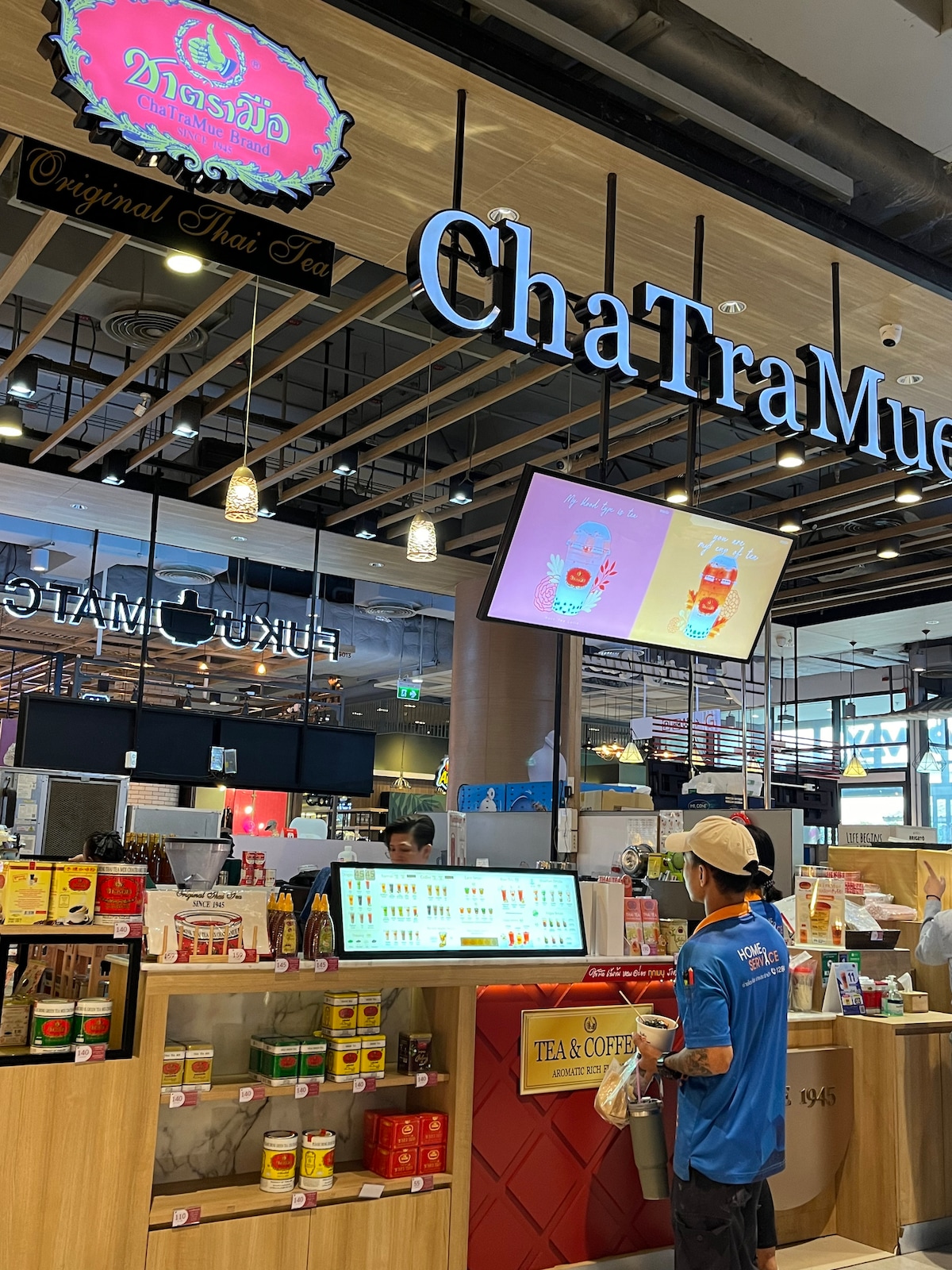 A vibrant tea and coffee shop is displayed, featuring a colorful menu illuminated on screens. Packages of various teas are arranged neatly on shelves, showcasing their branding. A staff member is engaged in service at the counter, contributing to the welcoming environment.
