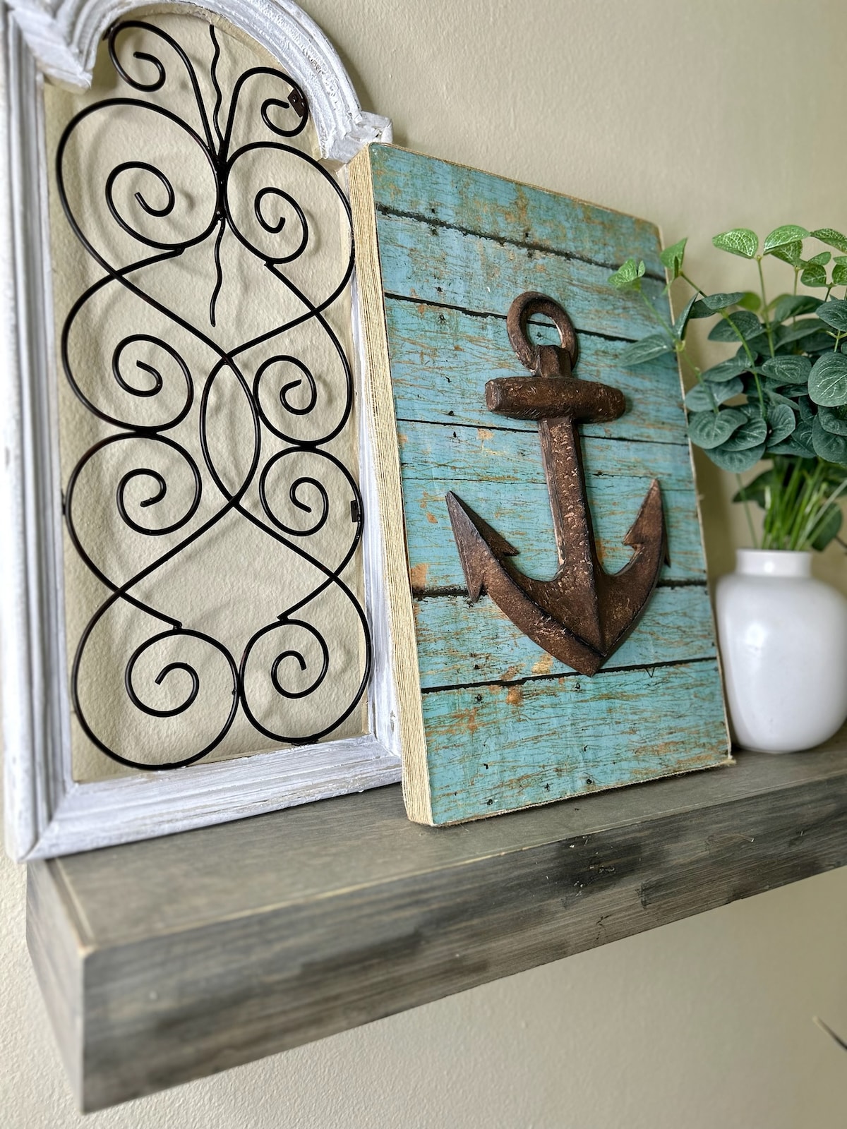 A decorative shelf displays an ornate metal design framed in white alongside a wooden piece featuring a rusted anchor against a turquoise background. A small white vase with greenery adds a touch of freshness to the composition.