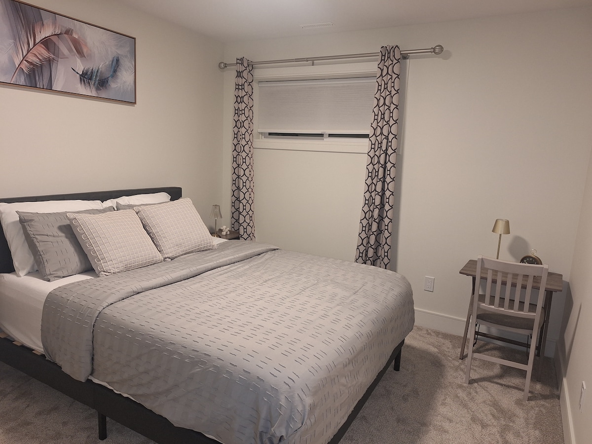 A spacious sleeping area features a comfortable bed with light grey bedding and decorative pillows. Minimalistic artwork hangs above the headboard. A small desk and chair are positioned in one corner, with light filtering in through a window adorned with patterned curtains.