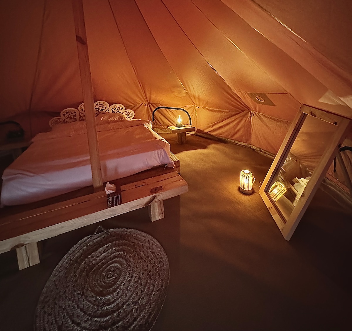 Inside a cozy glamping tent, a spacious bed rests on a wooden platform, complemented by soft lighting from candles. A large mirror and a woven rug are positioned nearby, enhancing the serene environment. The tent's fabric walls create a warm, inviting atmosphere.