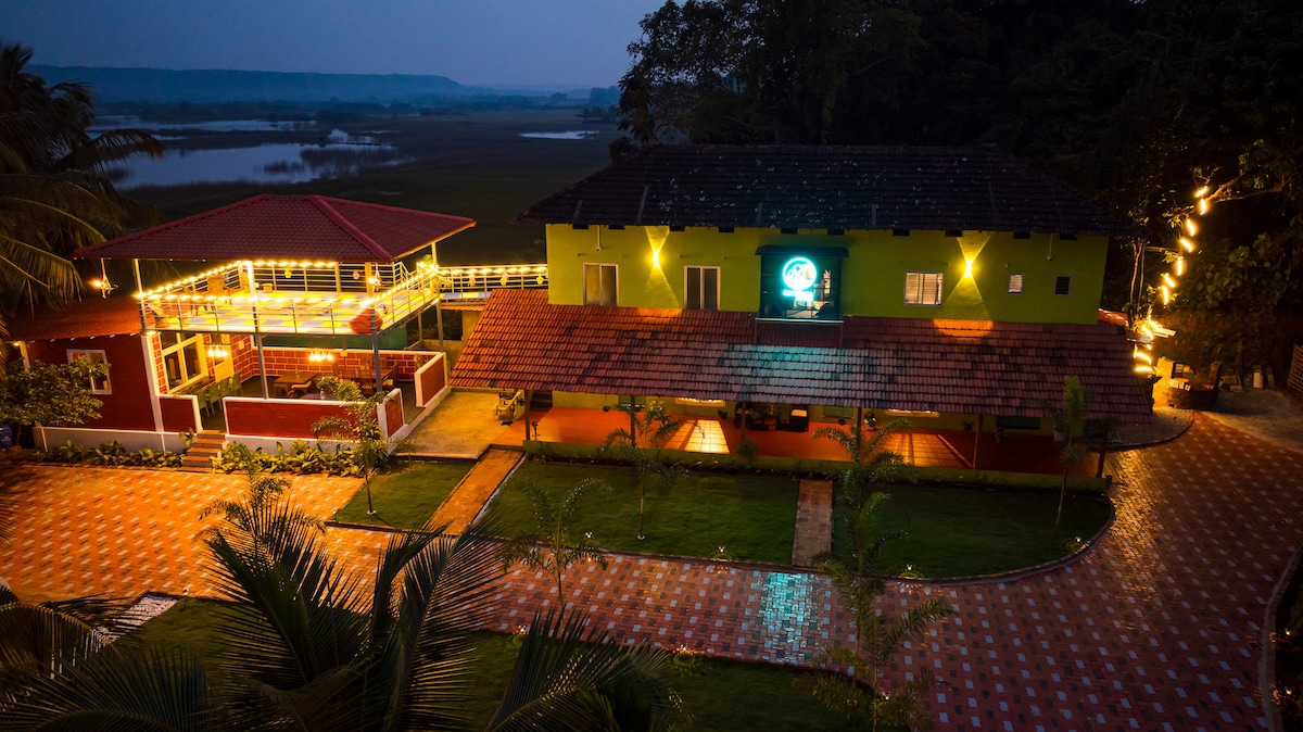 An aerial view of Cocobaba Farms captures the vibrant colors of the buildings, with warm lights illuminating the structures. The landscaped grounds feature a well-maintained pathway, surrounded by greenery, leading to a tranquil body of water in the background.