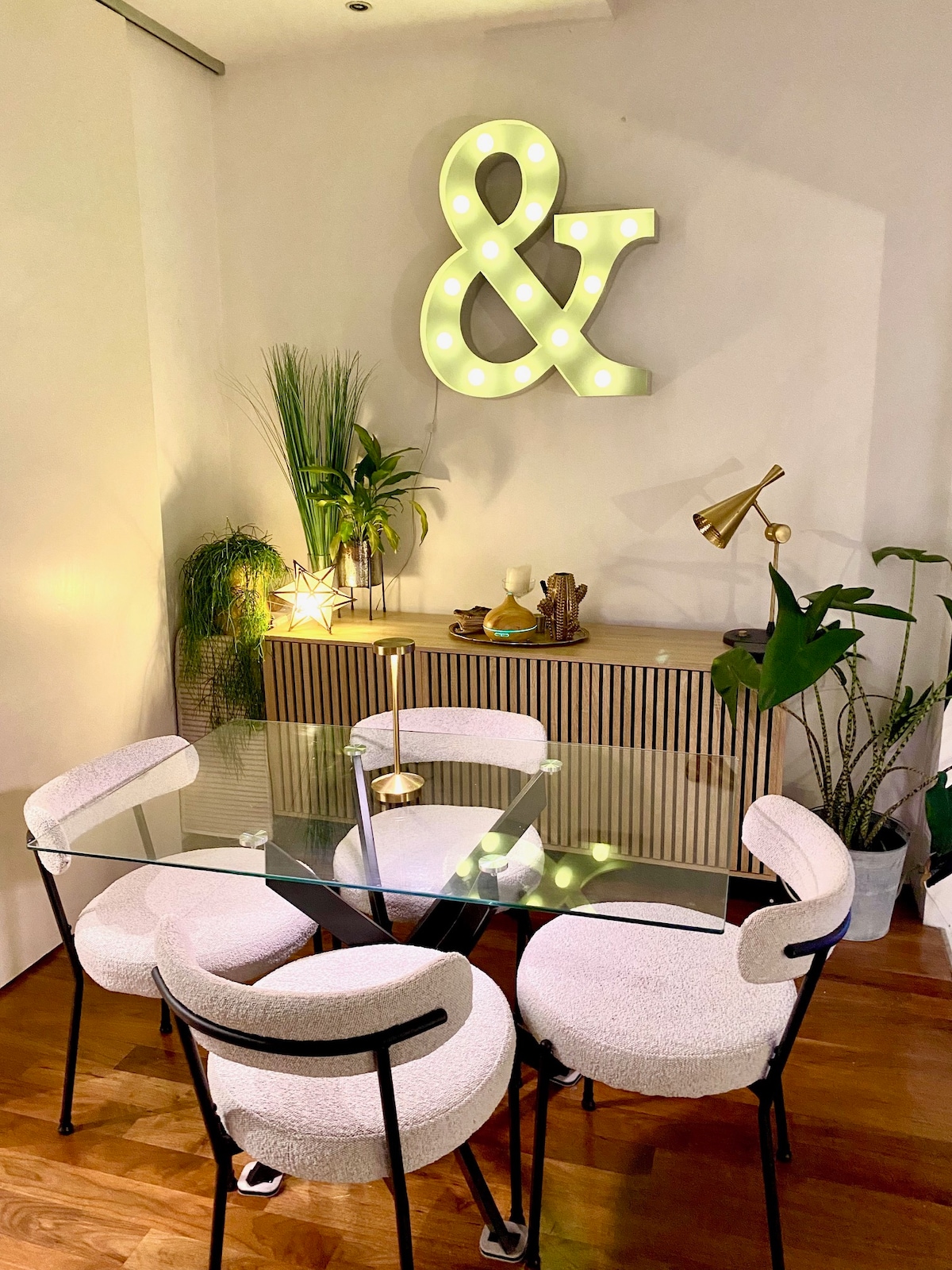 A contemporary dining area features a glass table surrounded by four textured chairs. A decorative sideboard displays plants and decor items. A large illuminated ampersand symbol hangs on the wall, adding a unique touch to the softly lit space.