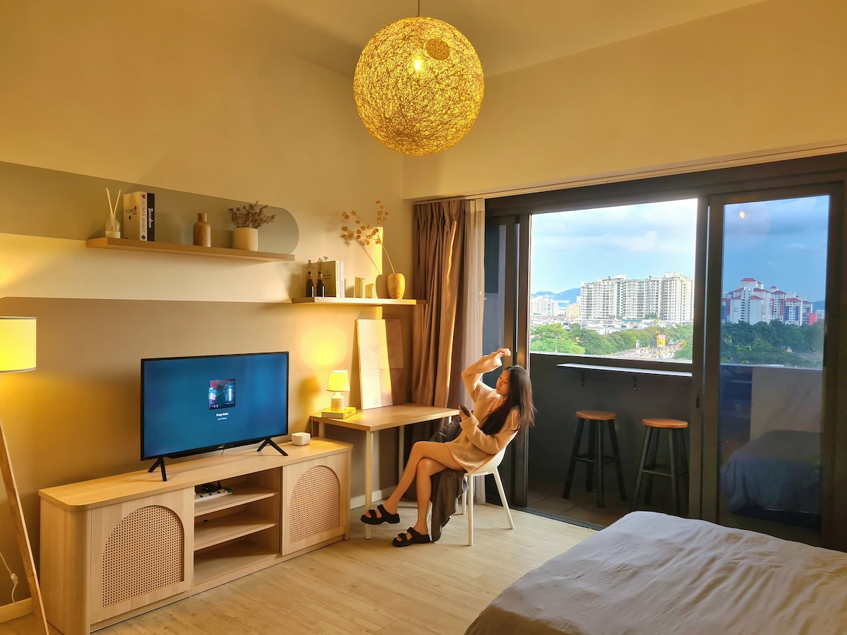 A well-lit studio showcases minimalist wooden accents and a cozy queen-size bed. A smart TV is positioned on a wooden unit, while a chair and table create a workspace. Large windows offer views of the city skyline, accompanied by natural light and a chic pendant lamp.