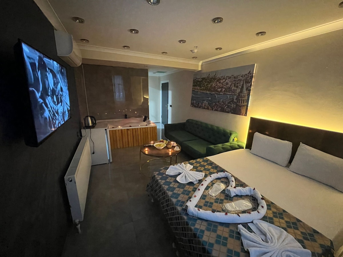 A well-appointed living space features a spacious bed adorned with a heart-shaped arrangement of towels. A green sofa complements the area, while a wall-mounted television and a kitchenette enhance convenience. Soft lighting highlights the modern decor, creating a comfortable environment.