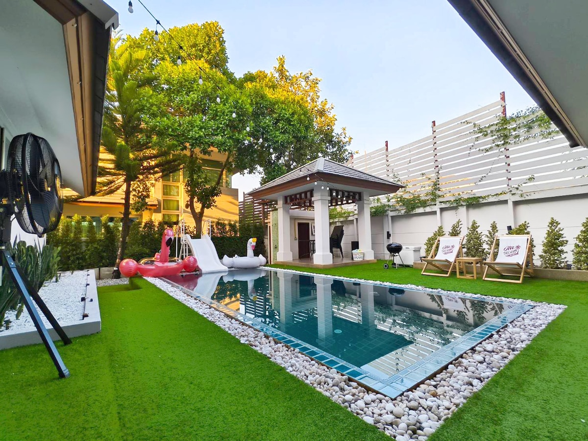 A private swimming pool is surrounded by lush green grass, featuring colorful inflatable slides for added fun. Sun loungers are positioned nearby, with a patio area visible in the background. Decorative stones outline the pool, creating a neat and inviting recreational space.
