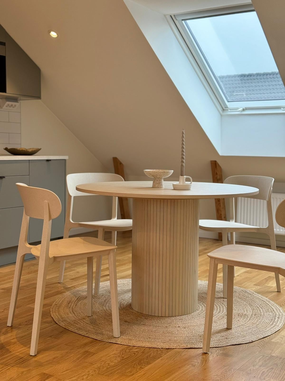 A dining area features a round table supported by a cylindrical base, surrounded by four light-colored chairs. A textured area rug is placed under the table, while a skylight allows natural light to fill the space.