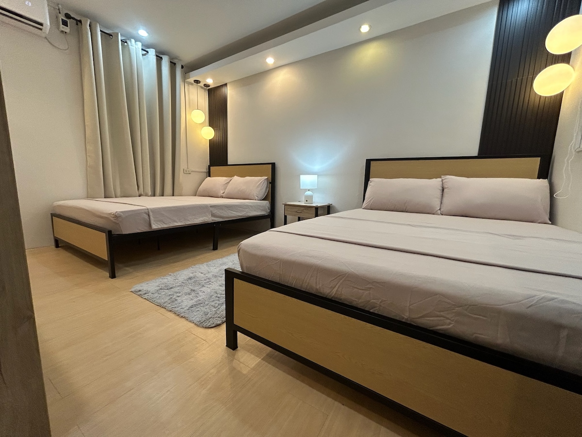 The bedroom features two spacious beds with neutral-toned linens. A small bedside table holds a lamp, providing soft light. Light-colored curtains frame the window, and a plush rug lies on the floor, enhancing the room's cozy feel.