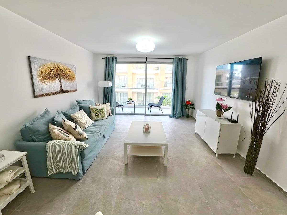 A modern living area features a light blue sofa adorned with various cushions. A simple coffee table sits in front, with a decorative item placed on it. Large windows dressed in sheer curtains allow natural light to fill the space, while a television is mounted on the wall.
