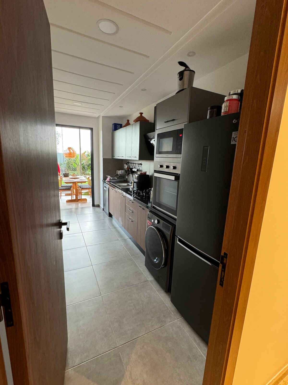 A narrow, modern kitchen reveals sleek cabinetry and appliances. A stylish refrigerator and stacked oven units stand on one side, while a sink and cooking area offer functionality. Light enters through a large window, illuminating the elegant tile flooring and providing a view of the outdoor seating area.