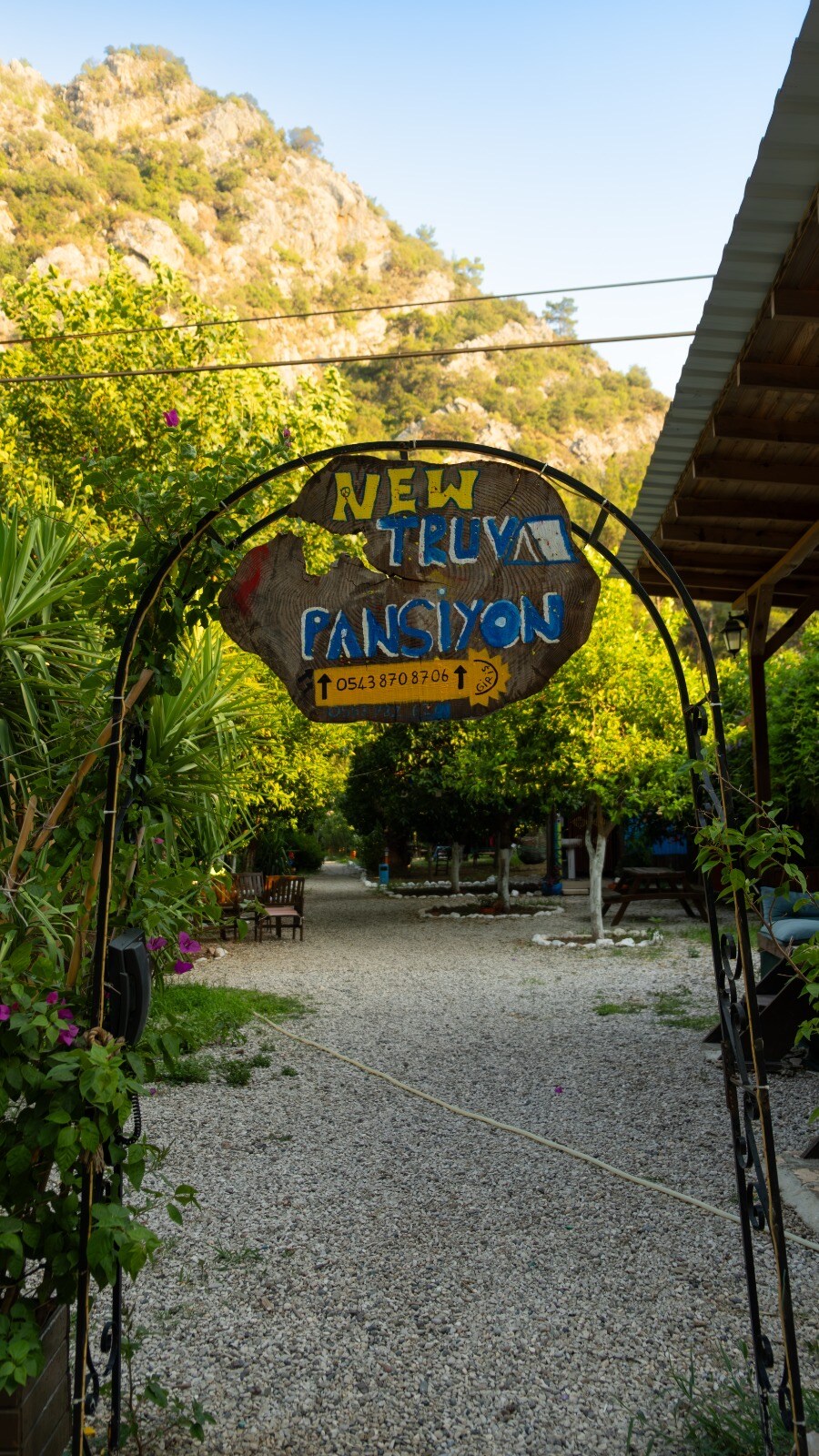 An arched wooden sign displaying 'NEW TRUVA PANSIYON' serves as an entrance marker. Surrounding greenery includes trees and flowering plants, framing a gravel pathway that leads into the area. Natural sunlight filters through the foliage, creating a serene environment.