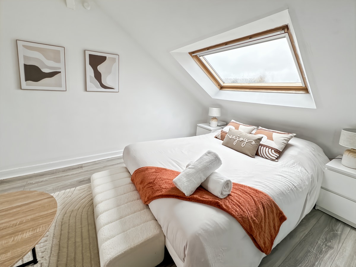 A well-lit sleeping area features a spacious bed with decorative pillows and a cozy blanket, complemented by a large window that allows natural light to enter. A bedside table and lamp accompany each side of the bed, while a circular rug adds warmth to the space.