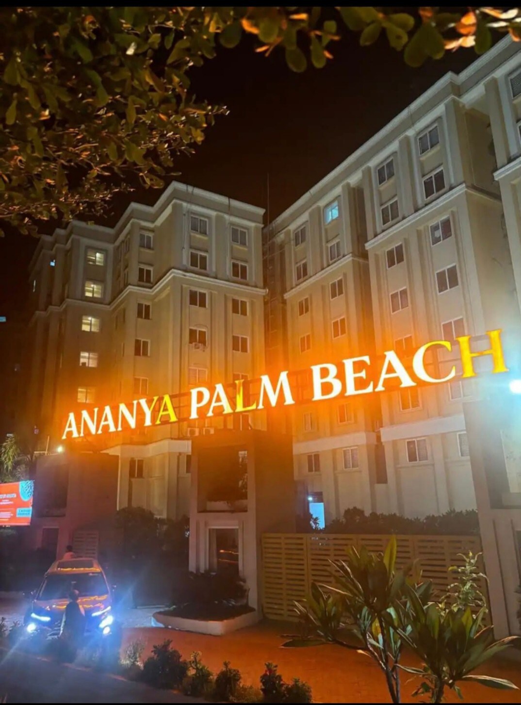 The illuminated sign for Ananya Palm Beach is prominently displayed at night, casting a warm glow over the entrance. The surrounding greenery enhances the inviting presence of the building, which is highlighted by soft exterior lighting.