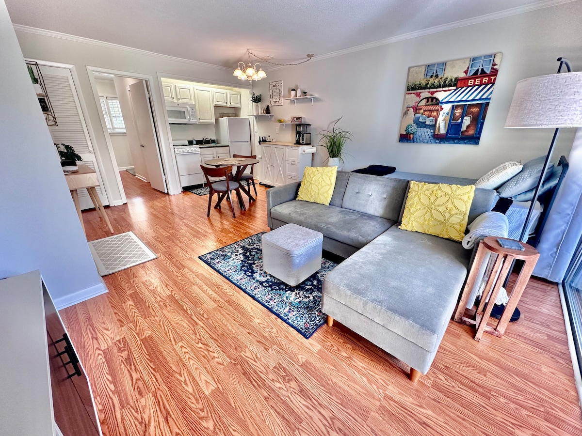 An open living space features a sectional sofa with yellow cushions and a decorative throw, paired with a plush ottoman. A small round dining table sits nearby. A kitchen area can be seen in the background, complemented by warm wood flooring and natural light from large windows.