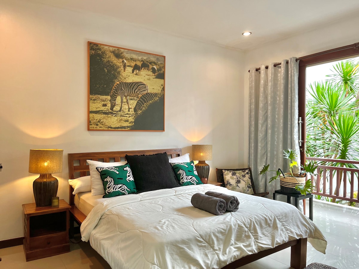 A cozy bedroom features a queen-size bed with decorative pillows and a soft blanket. Natural light filters through a window adorned with sheer curtains, highlighting a nature-themed artwork above the bed. Two bedside lamps provide gentle illumination, with additional greenery visible in the room.