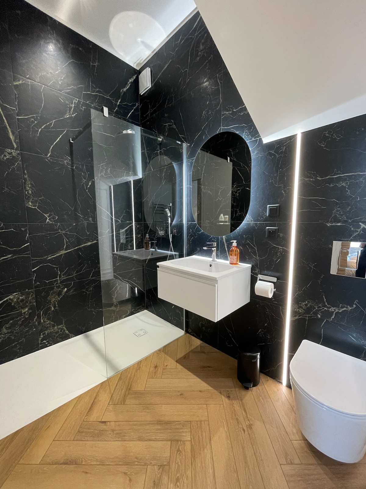 A modern bathroom features dark marble walls accented by a large mirror and stylish lighting. A suspended vanity offers storage beneath a circular mirror. The flooring is herringbone patterned wood, and a sleek toilet is positioned near the wall.