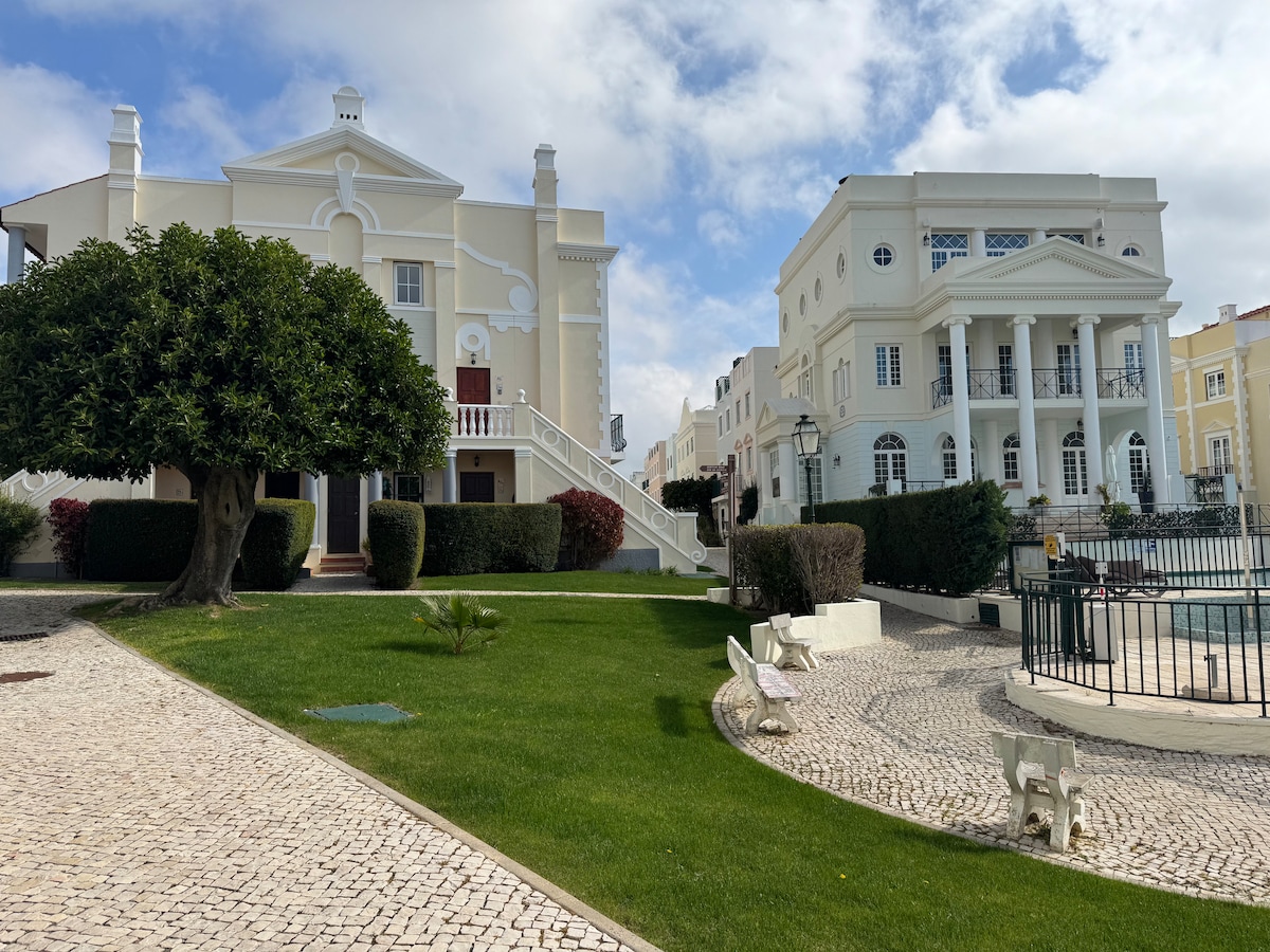 Two elegant buildings are framed by a well-maintained green lawn. One structure features classic architectural details with a grand staircase, while the other showcases neoclassical elements. Stone pathways and manicured hedges complement the surroundings, creating a serene atmosphere.