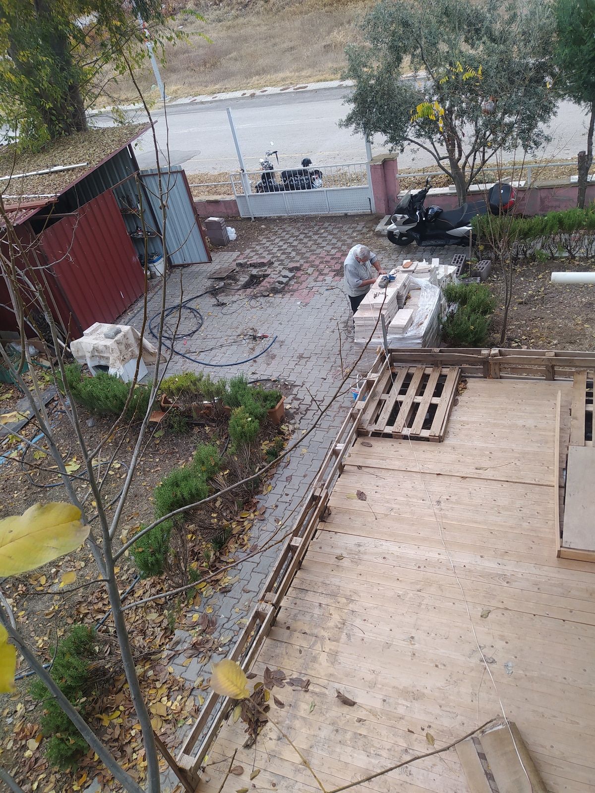 An outdoor area is visible, featuring a wooden deck and stone pathways. Various building materials, including pallets and bricks, are arranged at one side. A person is seen working at a table while a shed stands nearby. Trees and shrubs line the edges of the space.