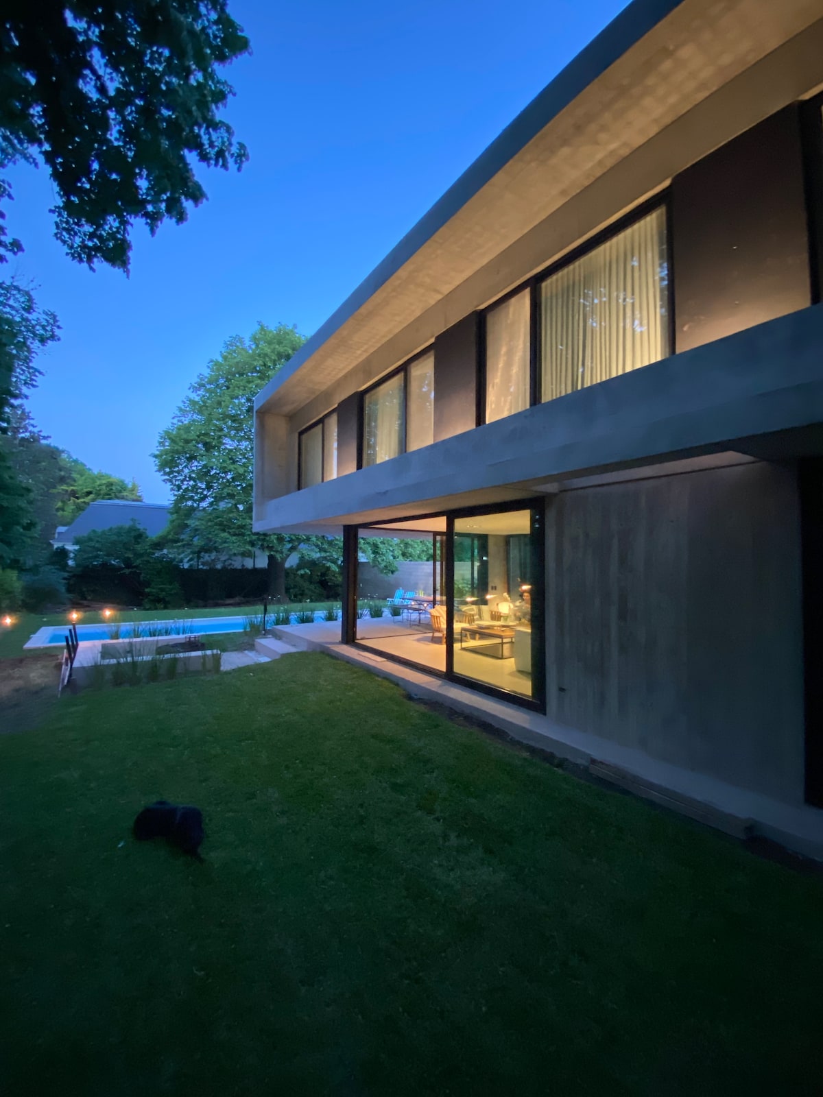 The exterior of a modern home is illuminated softly at dusk, showcasing large glass windows and a light-toned exterior. A landscaped area with well-maintained grass leads to a serene pool area in the background, surrounded by trees that add natural greenery.