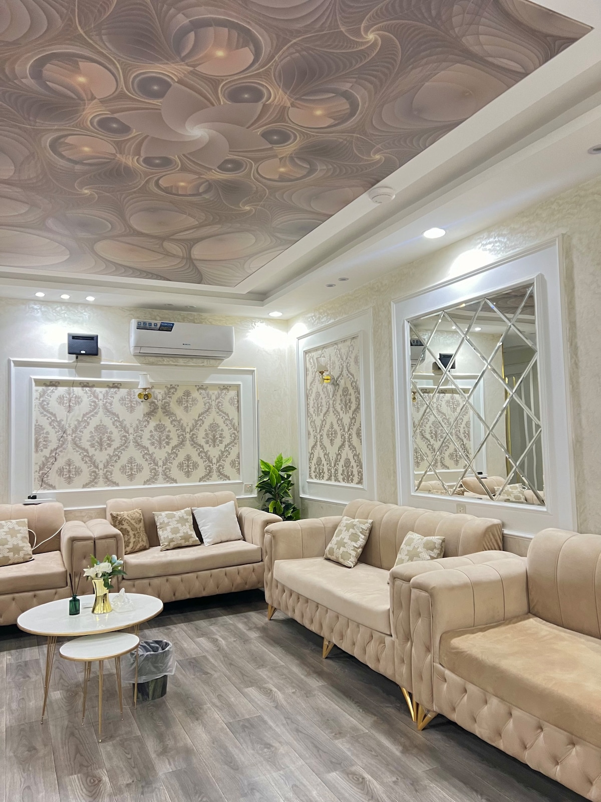 A stylish living area features light-toned, plush sofas arranged around a small, round coffee table. Elegant wallpaper adorns the walls, complemented by decorative mirrors. Soft lighting enhances the space, while a small potted plant adds a touch of greenery.