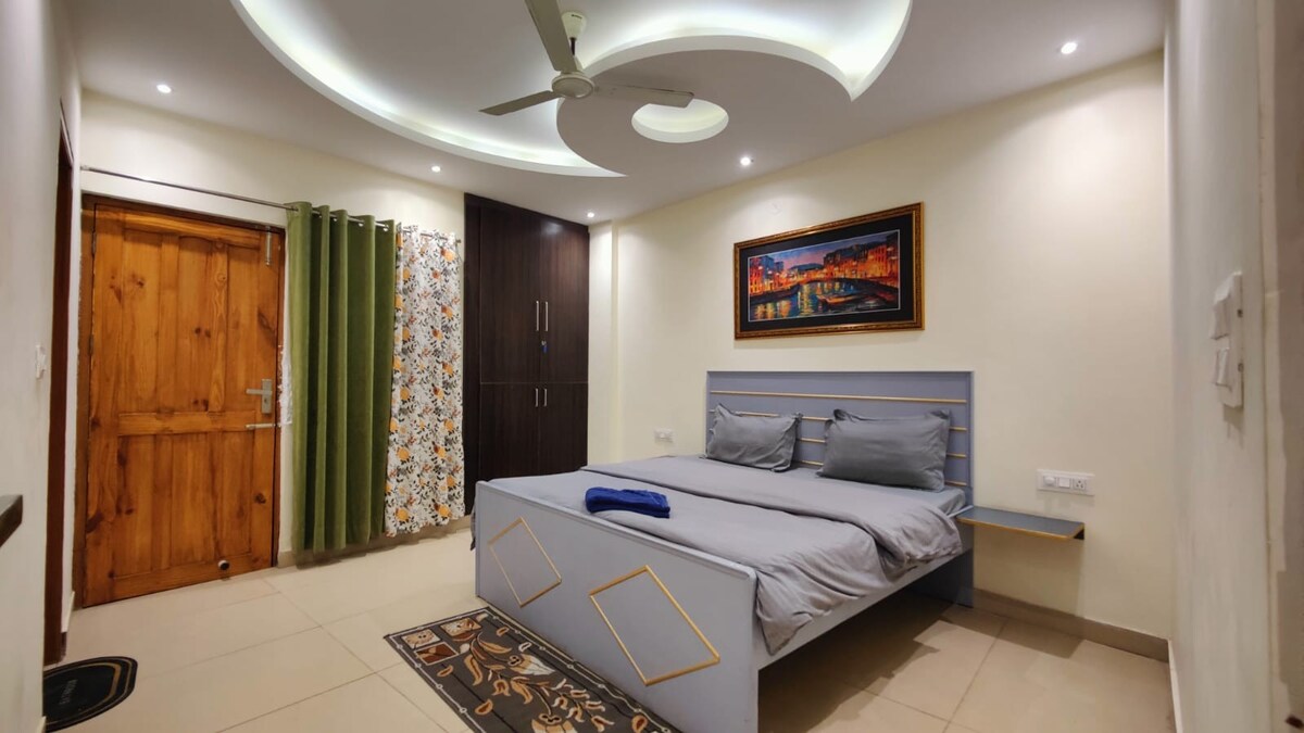 A modern bedroom features a spacious bed with neutral bedding, complemented by a decorative wall art piece. A ceiling fan is positioned above, and natural light enters through a window with green curtains. The entrance is highlighted by a wooden door and patterned rug.