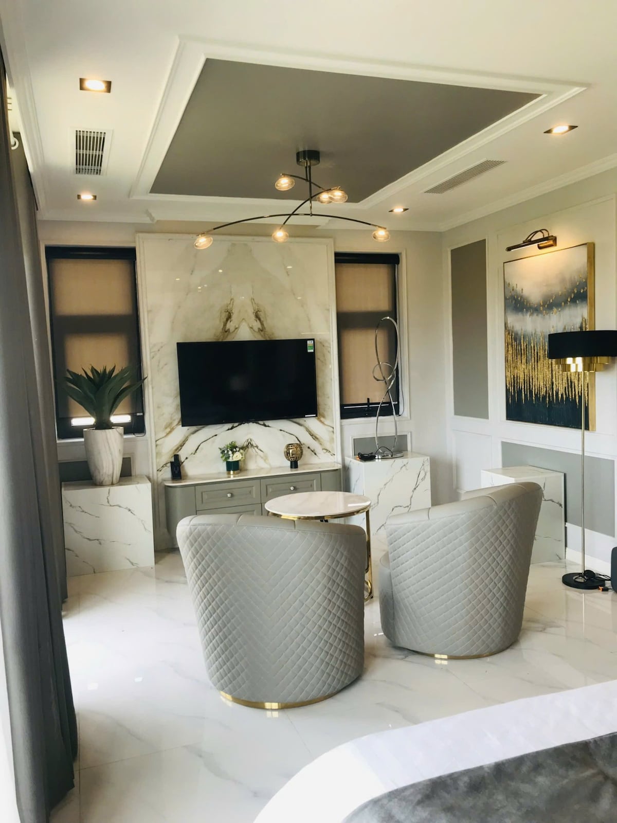 A stylish living area is presented with modern decor. Two textured armchairs are positioned near a sleek marble accent wall, which features a flat-screen television. A small round table complements the seating, while natural light enters through the windows, enhancing the elegant atmosphere.