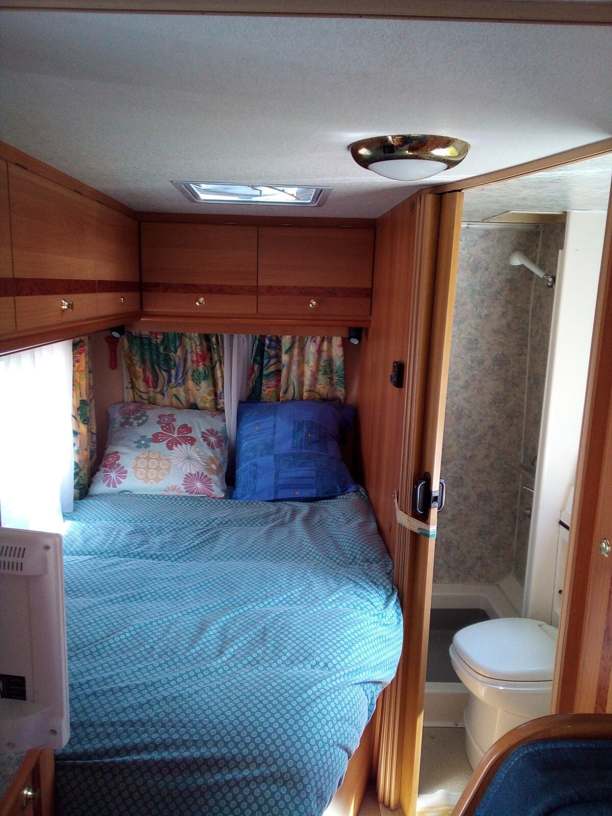 A compact sleeping area is furnished with a spacious bed covered in a blue blanket. Colorful patterned curtains frame the window, allowing natural light. A private bathroom area, equipped with a shower and toilet, is visible through an open doorway, emphasizing efficient use of space.