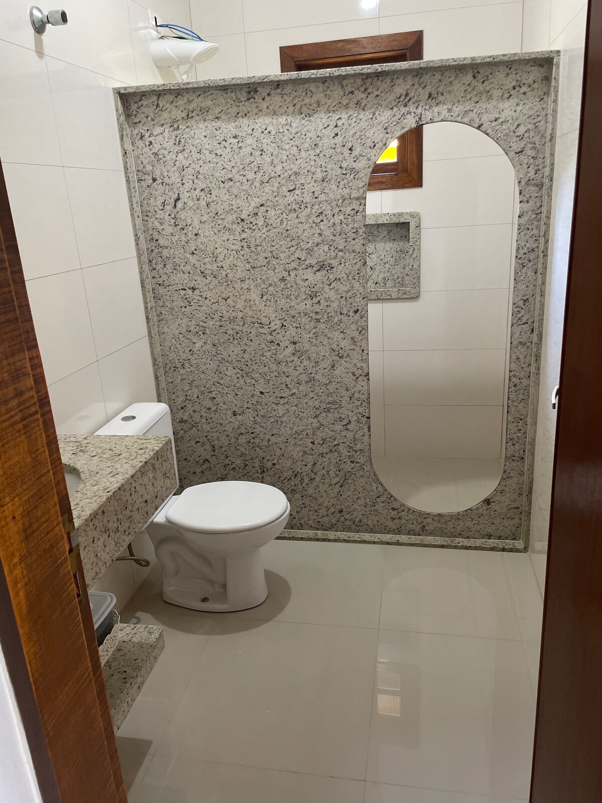 A modern bathroom features elegant granite wall finishes and a large oval mirror. A compact toilet and a sink with under-counter storage are included. Soft lighting enhances the clean and bright environment, with a window allowing natural light to enter.