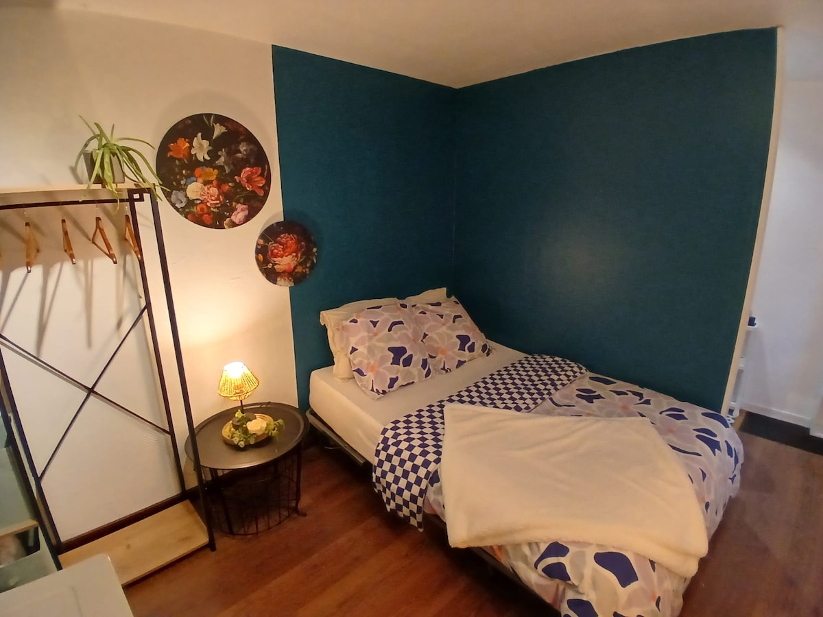 A cozy sleeping area is presented, featuring a bed with a floral-patterned duvet and a soft throw. A small bedside table holds a decorative plant and an illuminated lamp. The wall is adorned with floral art, and a clothing rack provides additional storage.