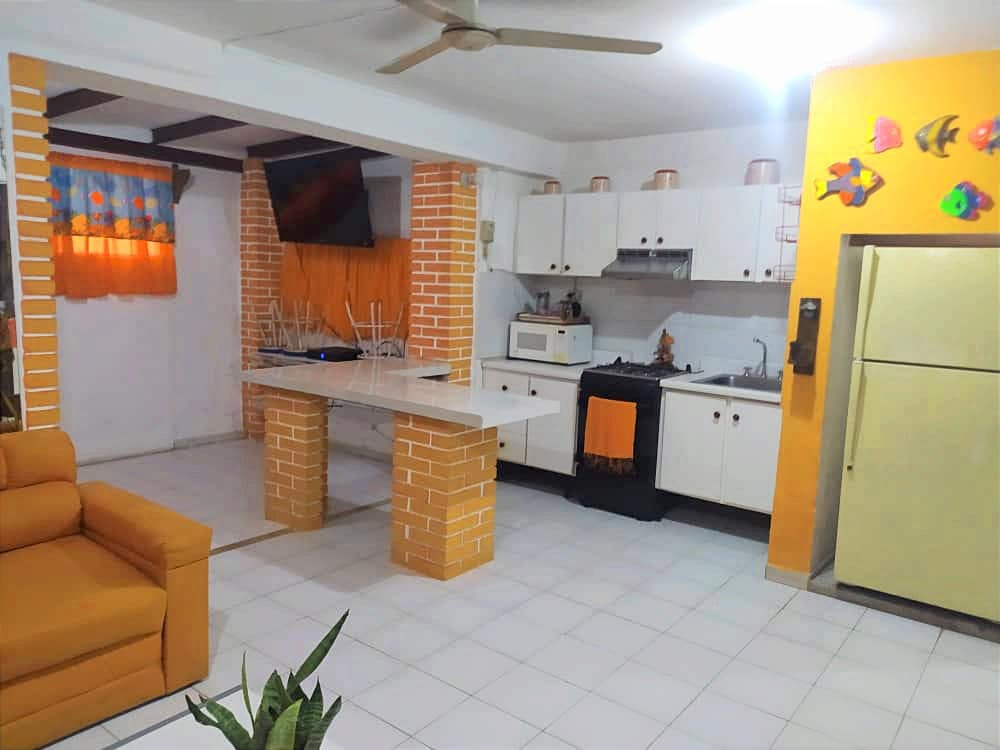 An open kitchen area features a bright orange accent wall and white cabinetry. A simple countertop separates the kitchen from the living space. A small dining area is visible with chairs stacked neatly. Natural light enters through a window draped with vibrant curtains.