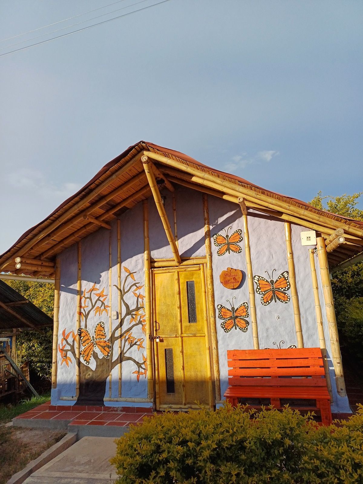 A charming cabin features a vibrant mural of butterflies and a tree, set against a light blue wall. A yellow door adds a pop of color, while a cozy orange bench is positioned outside. Lush greenery surrounds the structure, enhancing the natural setting.