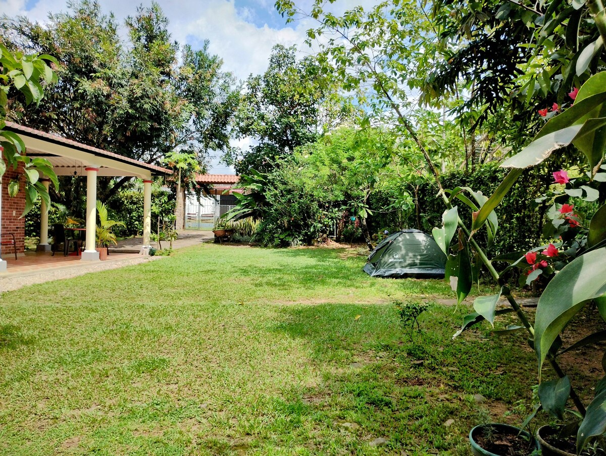 A spacious garden area is surrounded by lush greenery and trees. A tent is set up on the grass, while a patio is visible in the foreground, featuring columns and an open space for seating. Blooming flowers add colorful accents throughout the landscape.