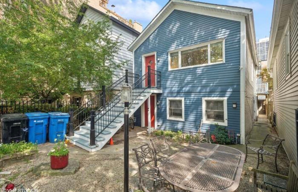 A blue two-story building is shown, featuring a staircase leading to the second floor entrance. The area is landscaped with greenery and flowers, alongside several outdoor trash bins. A round metal table with multiple chairs occupies a stone patio, inviting outdoor relaxation.
