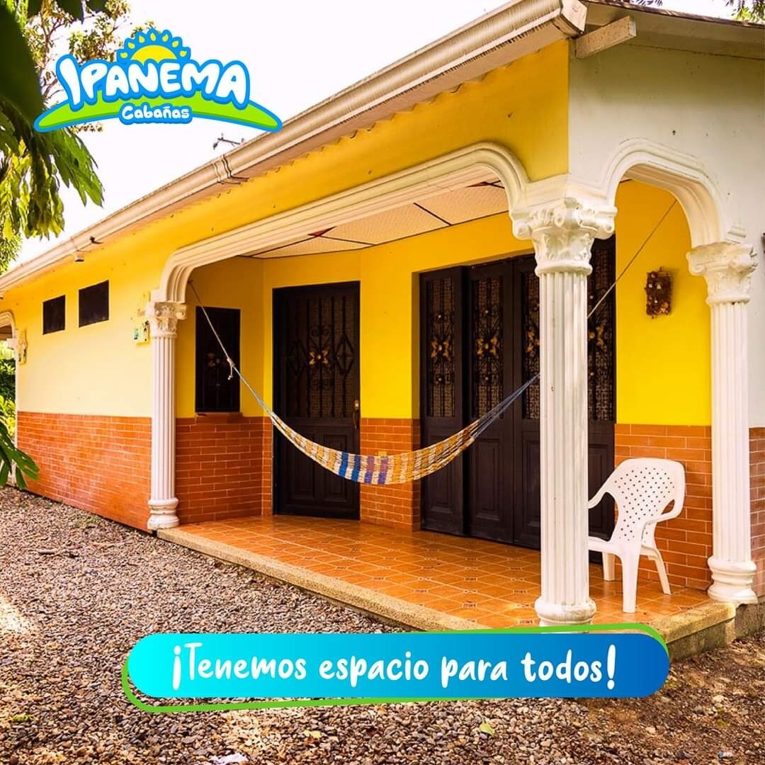 A welcoming entrance is highlighted by a patio featuring a colorful hammock and a white chair. The building boasts yellow walls and distinct columns, complemented by two sets of double doors and a neatly tiled floor. Surrounding greenery enhances the outdoor space.