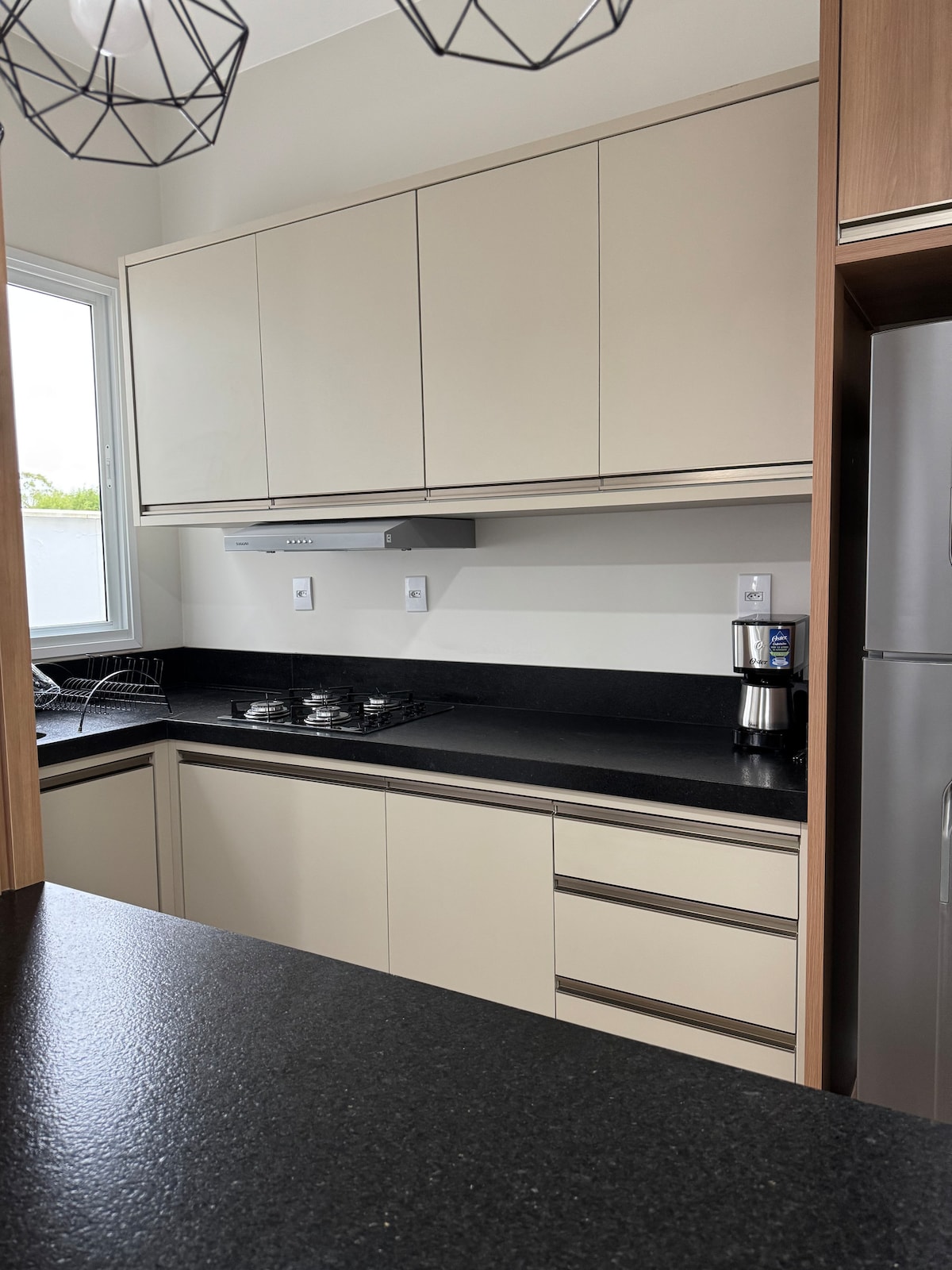A modern kitchen features sleek beige cabinetry and a black countertop. A gas stove and an espresso machine are located prominently, and natural light enters through a window, illuminating the space. A minimalist pendant light hangs above, adding to the contemporary design.