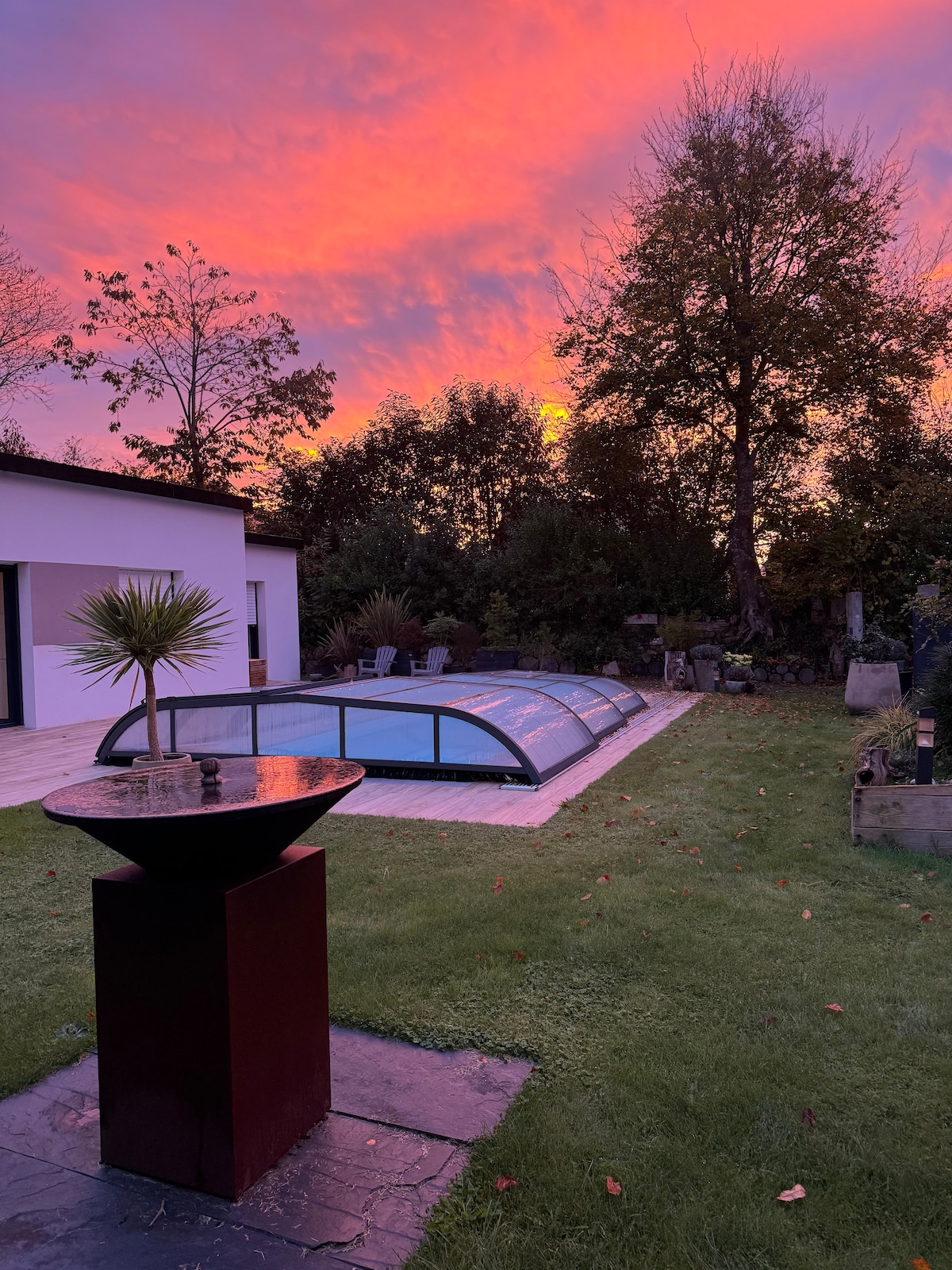 A serene outdoor area features a covered swimming pool reflecting the vibrant colors of a sunset. Lush greenery surrounds the space, with mature trees positioned in the background. A decorative water feature stands nearby, adding a tranquil element to the setting.