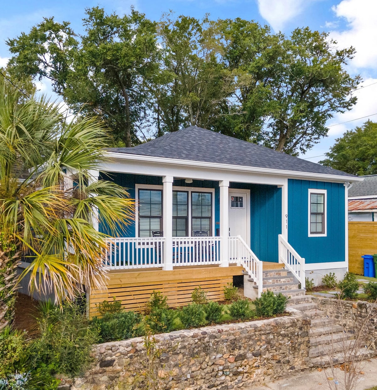The exterior of a renovated bungalow is showcased, featuring blue siding and a welcoming covered front porch. Lush greenery surrounds the home, with a set of steps leading to the inviting entrance. A stone pathway complements the vibrant landscaping.