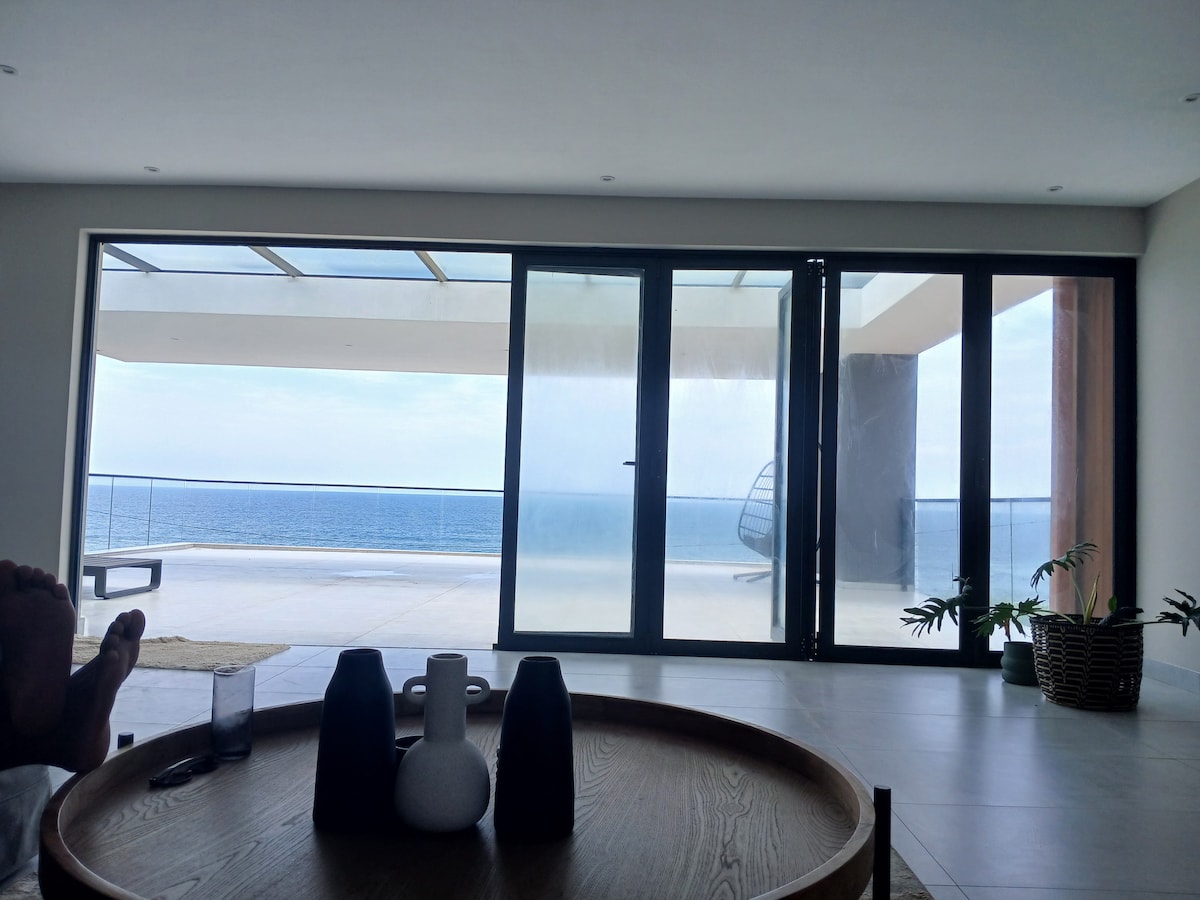 A spacious living area features large glass doors that open to an expansive view of the ocean. A circular wooden table with decorative items sits centrally, while a lounge chair and potted plants are visible in the foreground.